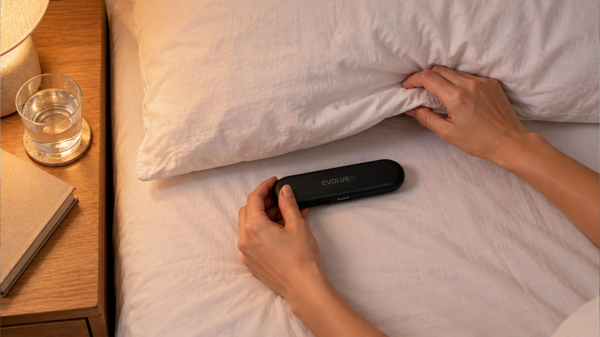 Ruce zvedají okraj polštáře a umisťují pod něj černý reproduktor XSleep.