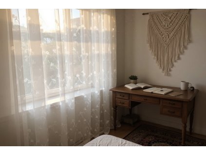 Sun drenched bohemian bedroom, morning light filtering through a sheer white floral embroidered curtain, illuminating dust motes dancing in the air; a woven