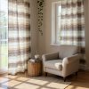 A cozy, sun drenched reading nook featuring floor to ceiling windows dressed with flowing, horizontally striped curtains in soft blues, browns, and crea