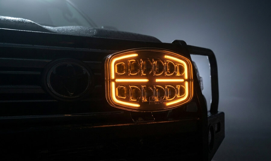 Részletes nézet az ovális kiegészítő távolsági LED fényszóróról kettős dinamikus helyzetjelzővel, aktivált narancssárga háttérvilágítással. A narancssárga vonal áthalad a fényszóró közepén, modern, dinamikus megjelenést kölcsönözve neki. - Kiegészítő távolsági LED fényszóró kettős dinamikus helyzetjelzővel - Ovális, 75W, 1500Lm, IP69K, R149, R148