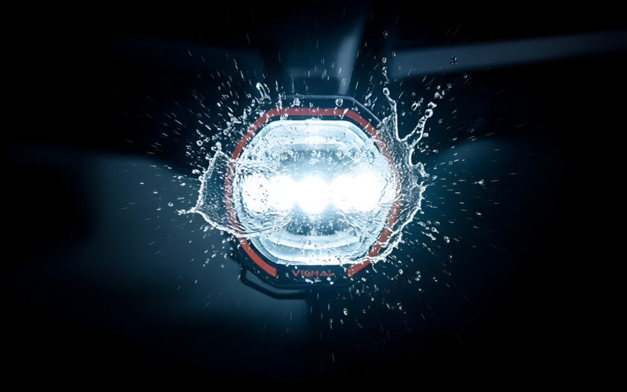 Detailní záběr na zadní stranu hliníkového těla přídavného 9palcového dálkového LED světla s kruhovým pozičním světlem. Husté chladicí žebrování zajišťuje, že se přídavné 9palcové dálkové LED světlo s kruhovým pozičním světlem nepřehřívá ani při dlouhé noční jízdě. - Přídavné dálkové LED světlo 9palcové s kruhovým pozičním světlem, 144W, IP68, R149, R10 Extrémní detailní záběr na kulaté 9palcové dálkové LED světlo namontované na nárazníku terénního vozu. Světlo je plně rozsvícené (studený bílý paprsek). Přes objektiv světlometu stříká proud vody a bláta, což vytváří dynamické kapky a odlesky ve světelném kuželu. Textura mokrého kovu, skla a bláta je hyperrealistická a ostrá. Pozadí je tmavé, rozostřené, v pohybu. Fotografie evokuje sílu, odolnost a certifikaci IP68 v praxi. Filmový, akční styl. - Přídavné dálkové LED světlo 9palcové s kruhovým pozičním světlem, 144W, IP68, R149, R10