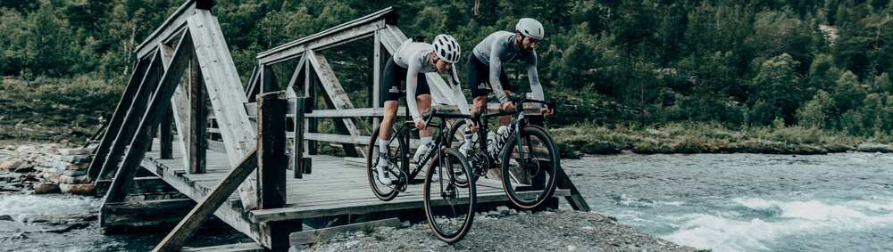 Dva cyklisté v oblečení Café du Cycliste přejíždějí dřevěný most přes řeku