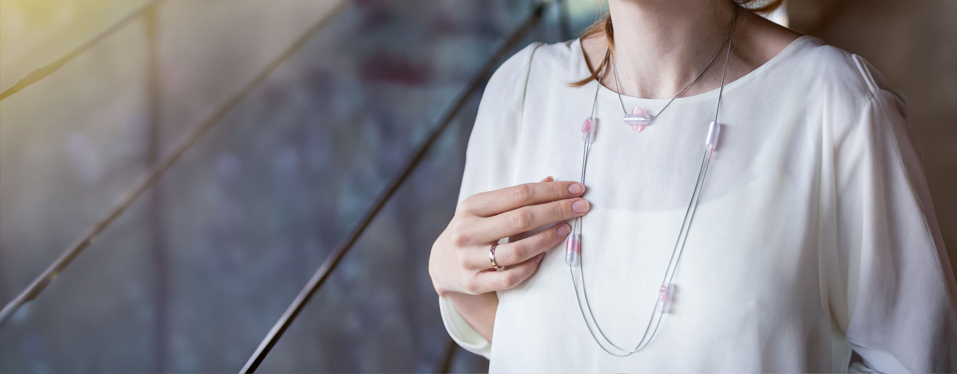 Detail ženy v bílé halence, která má na krku dlouhý minimalistický náhrdelník z tenkých kovových lanek s průsvitnými růžovými korálky; žena si jemně přidržuje náhrdelník rukou, v pozadí je rozostřené interiérové prostředí s oknem.