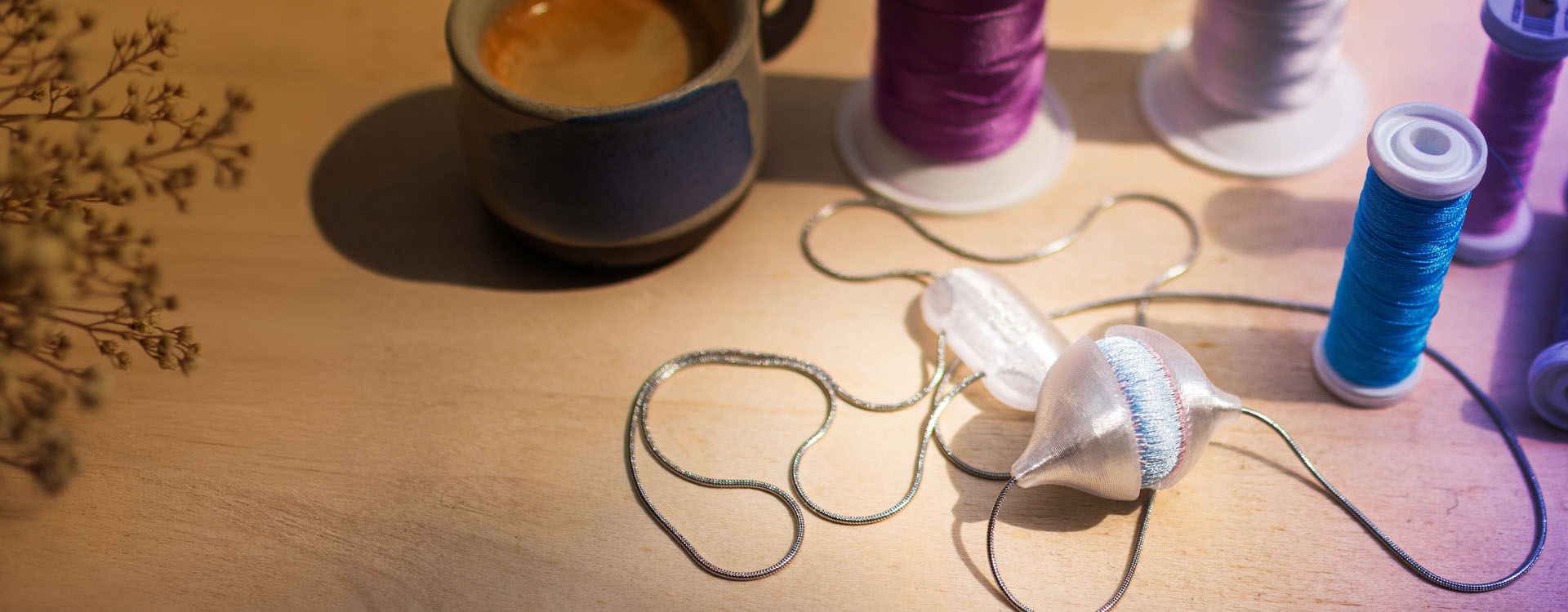 Zátiší na dřevěném stole s několika cívkami barevných nití (bílá, fialová, tyrkysová, šedá a růžová), jemným řetízkem a textilním přívěskem uprostřed; vpravo stojí malý keramický hrnek s kávou, vše nasvícené přirozeným světlem.