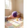 Close up of a handcrafted, purple toned crocheted juggling ball resting in the palm of a child's hand in a bright, playful nursery Sunlight streams in through a window casting a soft glow on a cheerful, pastel colo