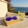 A sun drenched balcony overlooking the turquoise Mediterranean Sea, whitewashed walls adorned with bougainvillea cascading down A pair of purple heart shaped sunglasses rests carelessly on a stack of vintage postca