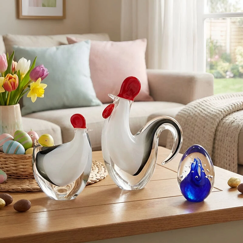 Velikonoční skleněné dekorace z českého skla - průhledný kohout a slepice s červenými detaily, umístěné na dřevěném stole s jarními tulipány, narcisy, velikonočními vajíčky a modré skleněné těžítko, vše v útulném domácím prostředí