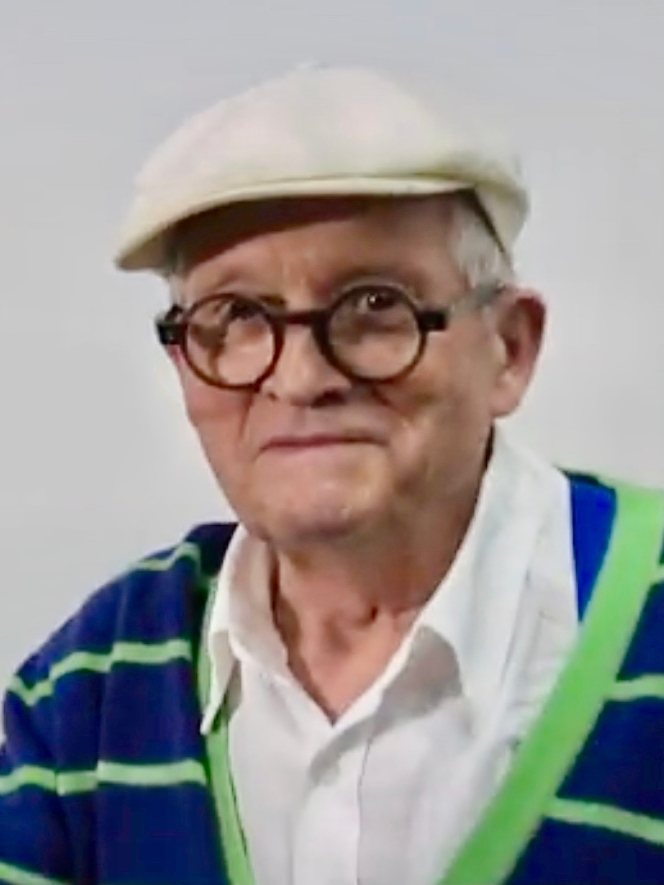 David Hockney v roce 2017 – starší muž v kulatých brýlích, světlé čepici a modře pruhovaném svetru