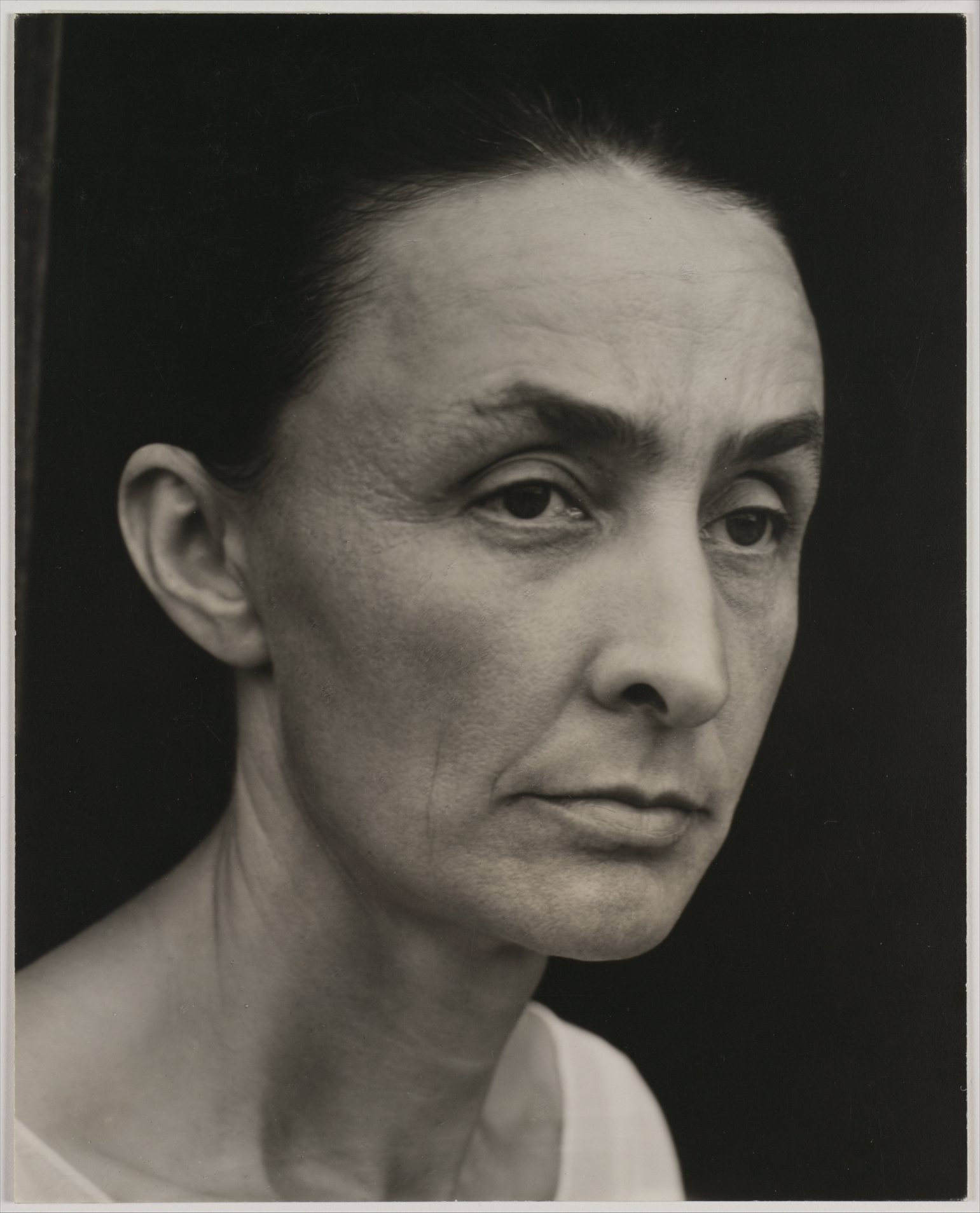 Černobílá fotografie Georgie O’Keeffe z roku 1932 od Alfreda Stieglitze – žena s přísným výrazem a staženými vlasy hledí do objektivu