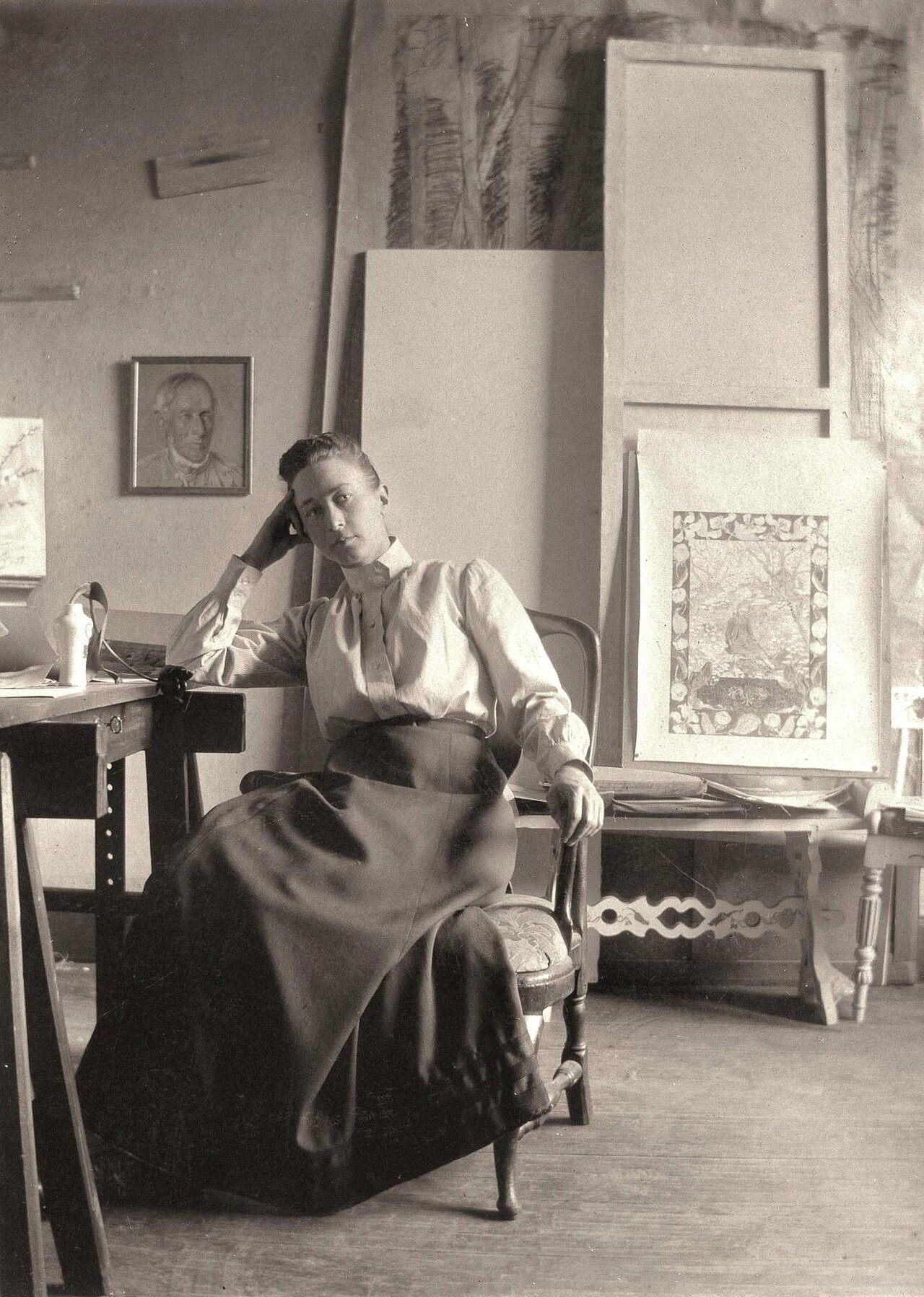 Černobílá fotografie Hilmy af Klint v ateliéru – žena v bílé halence a tmavé sukni sedí v křesle, za ní stojí obrazy a kresby