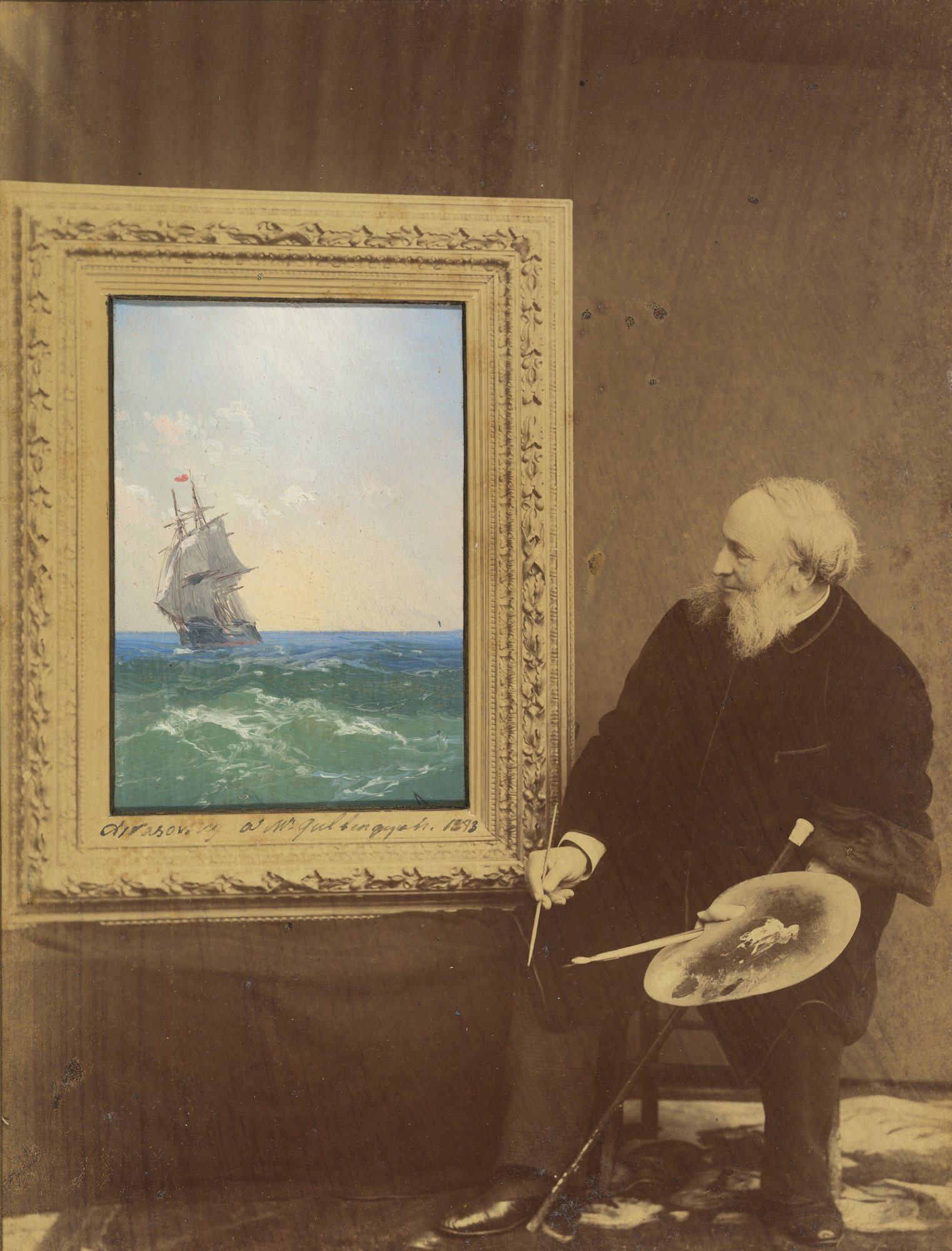 Fotografie Ivana Ajvazovského z roku 1893 – starý vousatý malíř s paletou sedí u stojanu s malým obrazem plachetnice na rozbouřeném moři