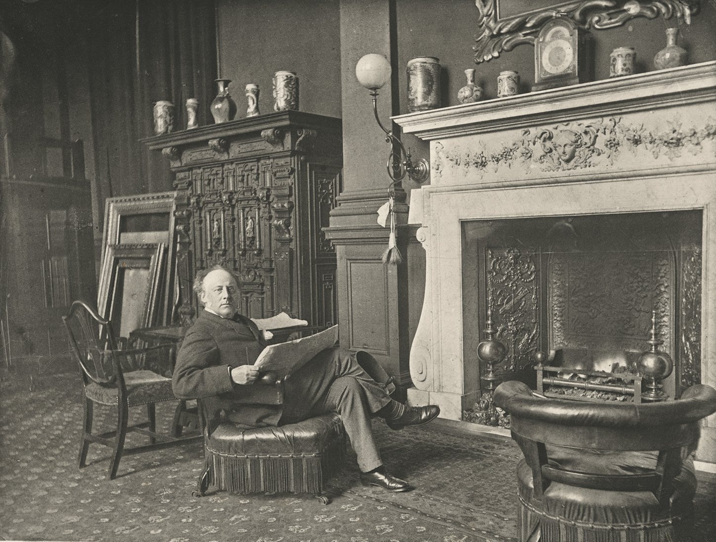 John Everett Millais na fotografii vydané roku 1884 – muž v tmavém obleku sedí s novinami v křesle u mramorového krbu ve svém londýnském ateliéru