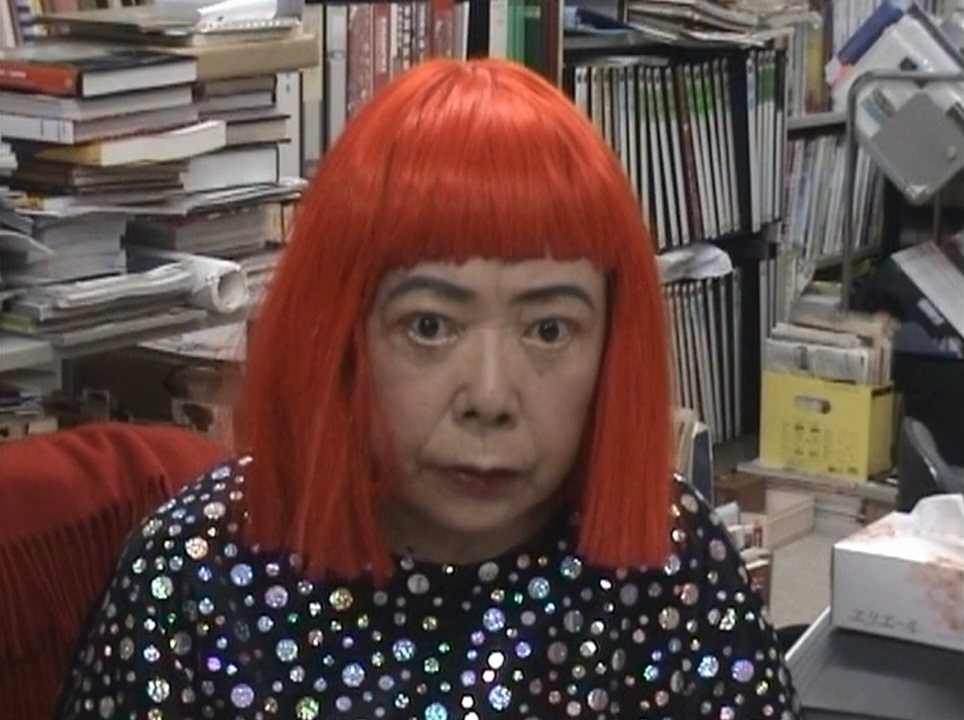 Yayoi Kusama v roce 2005 – žena s jasně červenou mikádovou parukou v puntíkovaných šatech v ateliéru plném knih