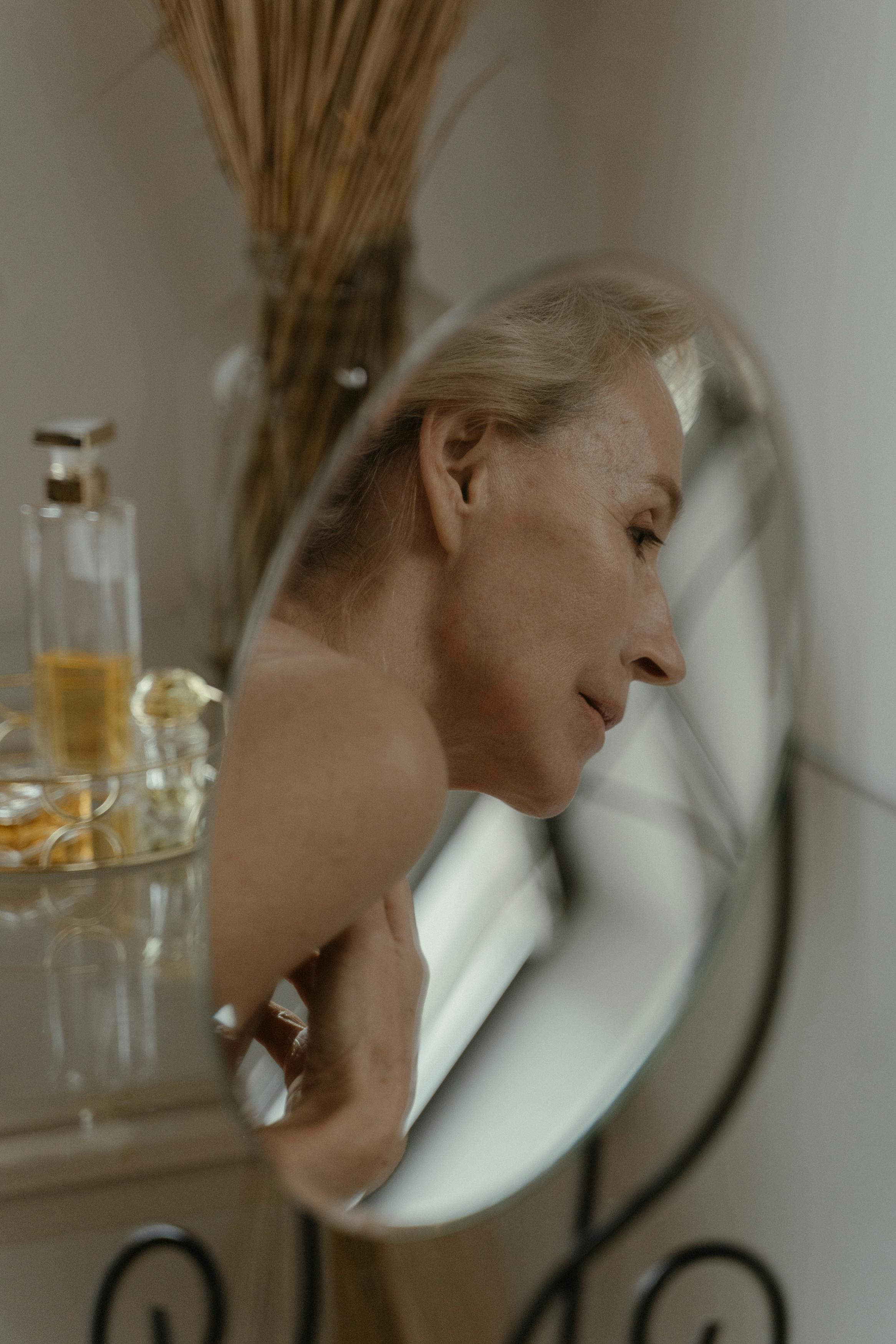 Žena se dívá do zrcadla v koupelně a věnuje si péči o tělo s láskou