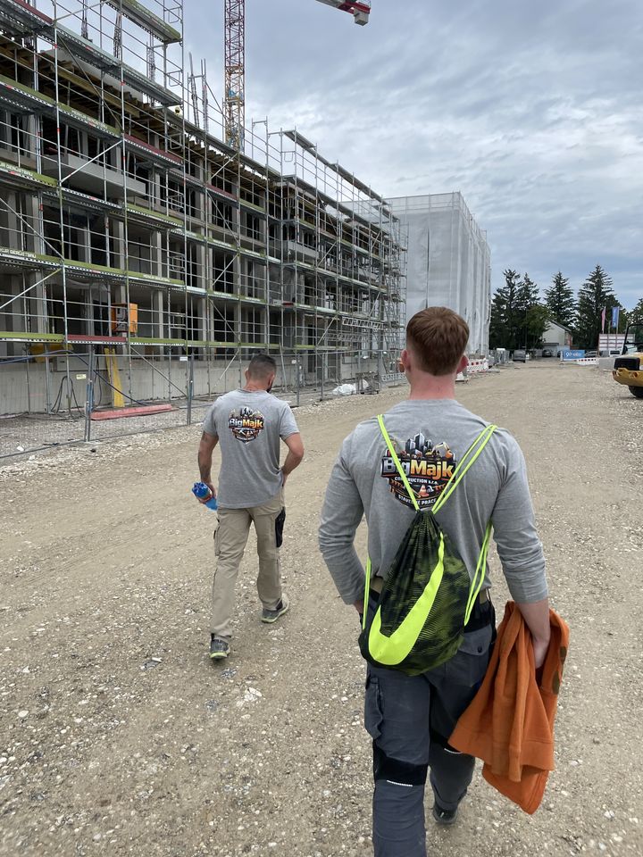 Dvaja pracovníci BigMajk Construction v sivých tričkách s potlačeným logom pred rozostavanou budovou obostavanou lešením