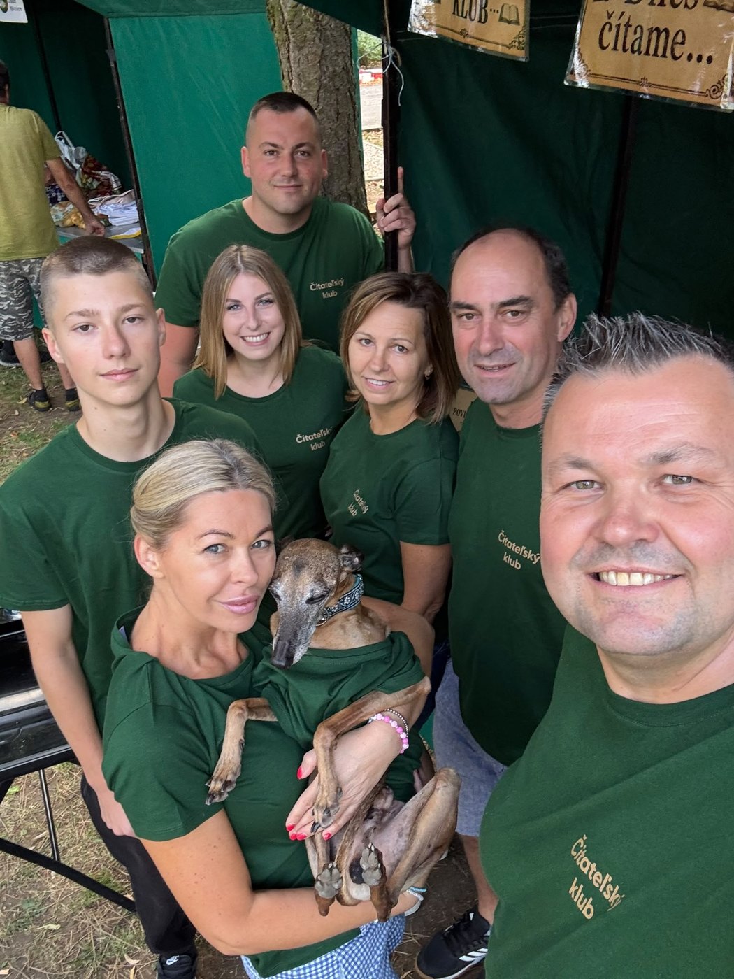 Skupinová selfie tímu v zelených tričkách so psom pod stanom