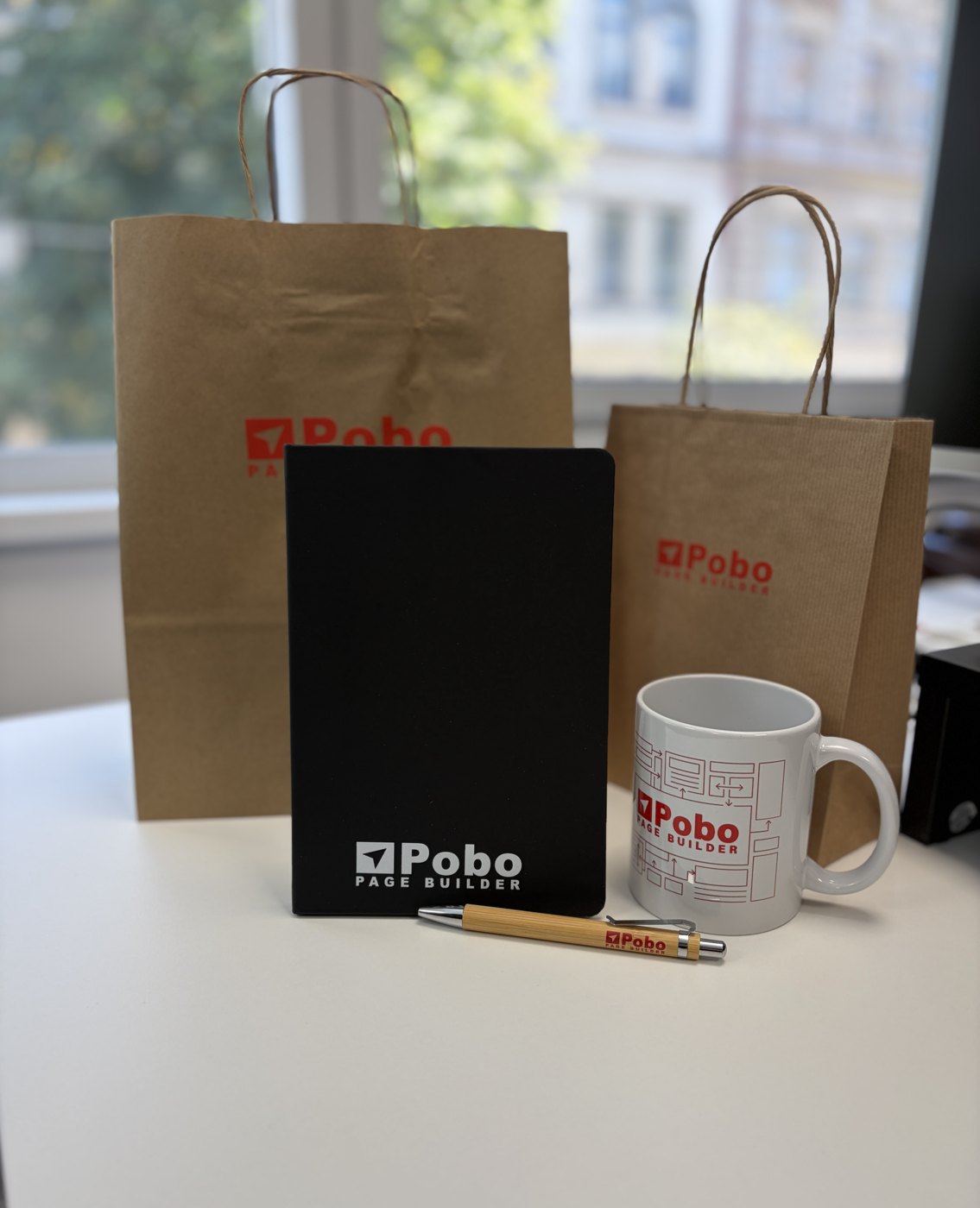 Papierové tašky, čierny zápisník, pero a hrnček s logom Pobo na stole