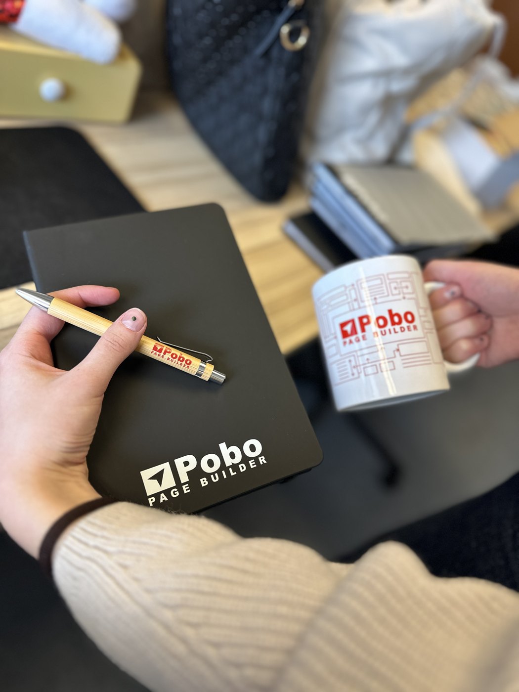 Ruky držiace čierny zápisník, drevené pero a hrnček s logom Pobo