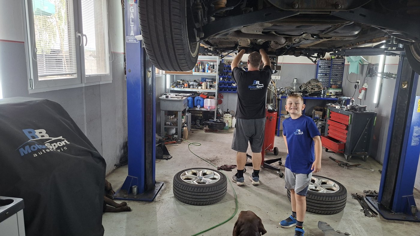 Mechanik pracuje pod vozidlom na zdviháku, vedľa stojí usmiaty chlapec