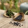 La Plantation – zelený a černý kampotský pepř v ratanových mističkách na dřevěném stole, zeleň v pozadí