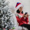 Žena v červeno-modrej mikulášskej čiapke sedí pri vianočnom stromčeku – moderná verzia klasickej sviatočnej čiapky