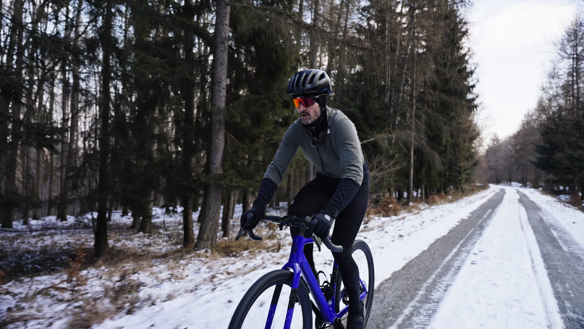 Jazdec v šedozelenej bunde na modrom gravel bicykli smeruje k nám po zasneženej lesnej ceste.