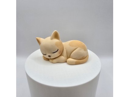 Figúrka spiacej krémovej mačky s oranžovými fľakmi na bielom podstavci.