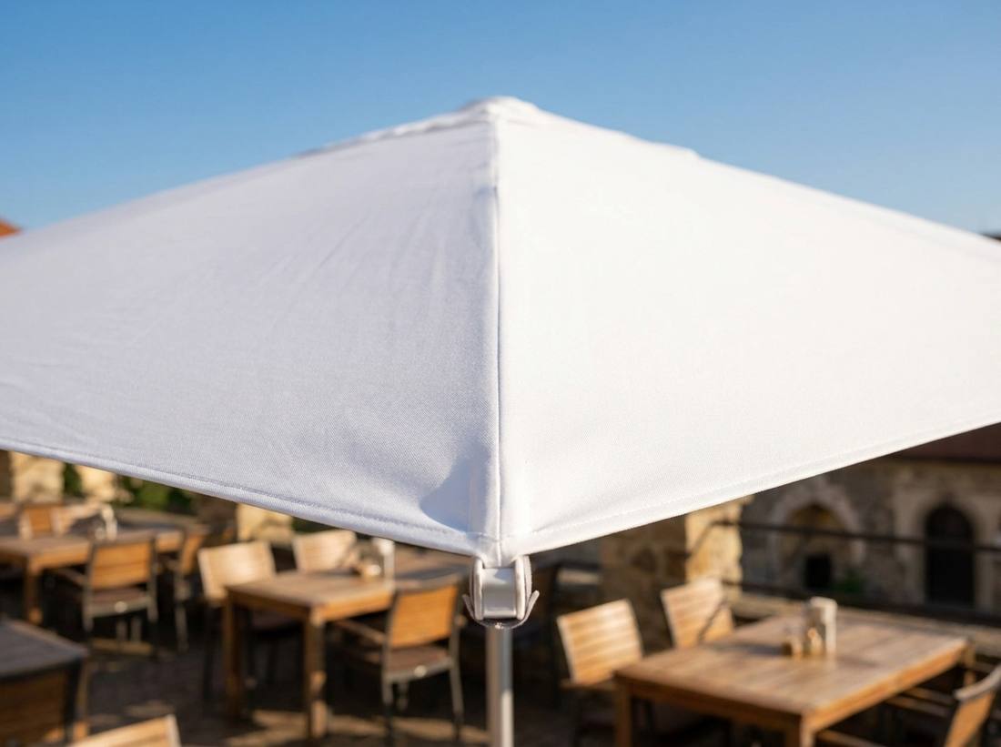 Detail rohu bílého potahu slunečníku Alu-Expert s koncovkou ramene nad restaurační terasou