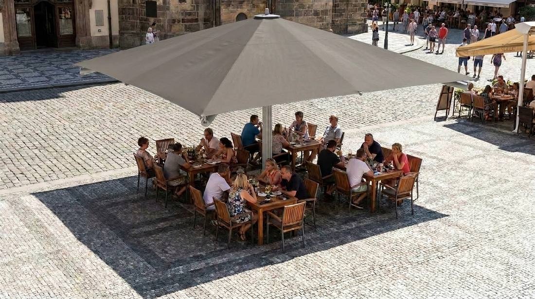 Pohled shora na slunečník Goliath 400 x 400 cm v béžové barvě zastiňující několik obsazených stolů