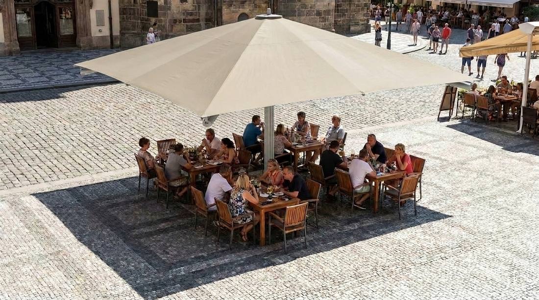 Pohled shora na slunečník Goliath 400 x 400 cm v světle béžové barvě zastiňující několik obsazených stolů
