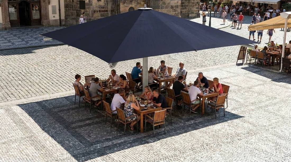 Pohled shora na slunečník Goliath 500 x 500 cm v modré barvě zastiňující několik obsazených stolů