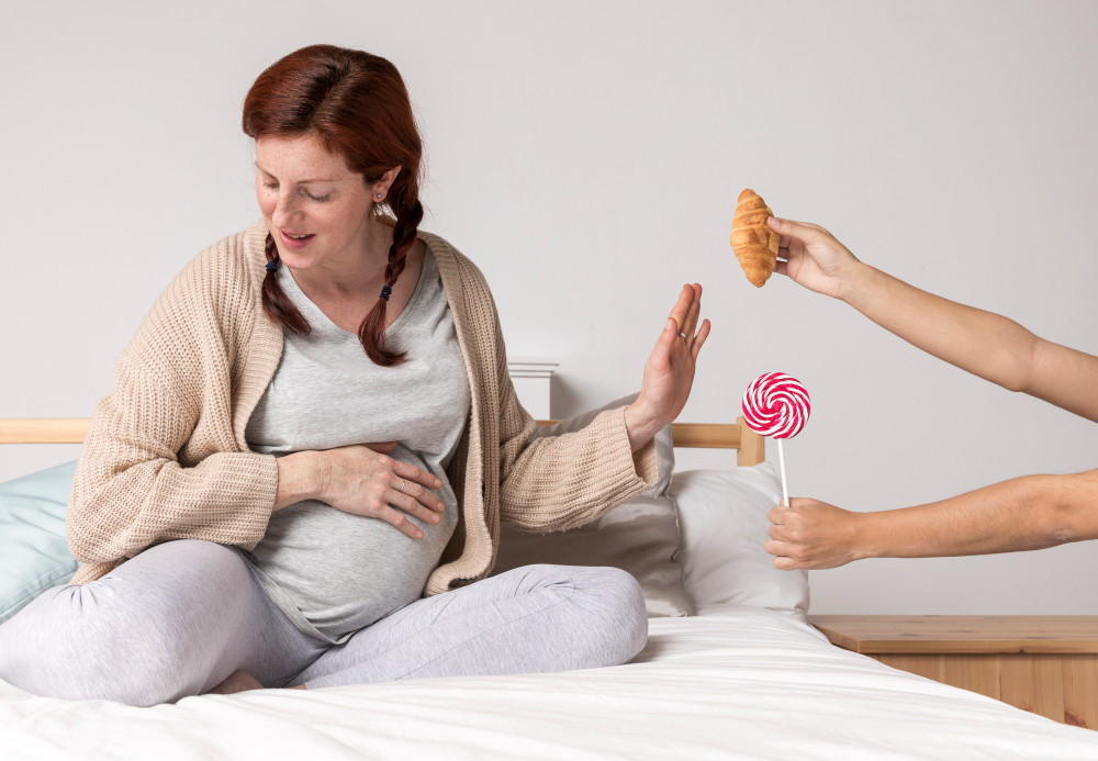 Těhotná žena sedící na posteli, drží si břicho a odmítá croissant a lízátko, které jí někdo podává – ilustrace k tématu těhotenská cukrovka hodnoty 5,2; 5,4; 5,5 a nutnost omezovat sladkosti