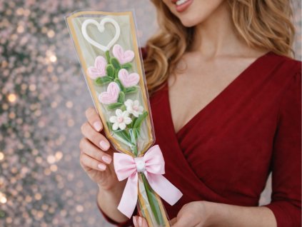 Žena v červených šatech drží mini kytičku z růžových srdíček a bílých kvítků vyrobenou z modelovacích drátků, dárkově zabalenou s růžovou mašlí.