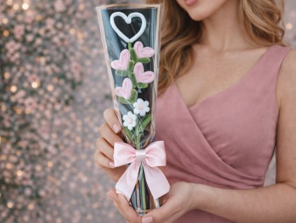 Žena v růžových šatech drží valentýnskou mini kytičku s růžovými srdíčky z modelovacích drátků v elegantním černém dárkovém obalu s mašlí.