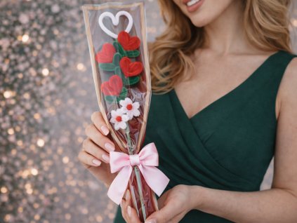 Žena v tmavě zelených šatech drží valentýnskou mini kytičku se čtyřmi červenými srdíčky a bílými kvítky z modelovacích drátků v dárkovém balení.
