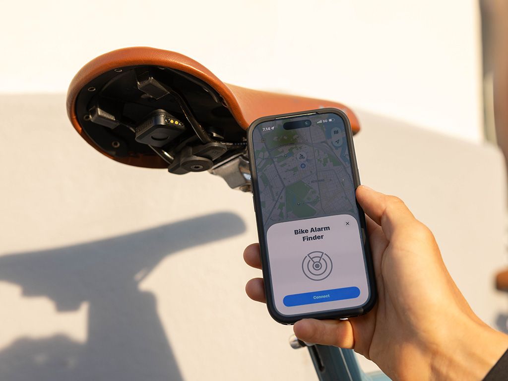 iPhone v ruce s aplikací Find My a nabídkou připojit Bike Alarm Finder, vedle alarm Quad Lock Bike Alarm and Finder (prodává se zvlášť) na adaptéru Quad Lock Saddle Rail Adaptor pod hnědým sedlem