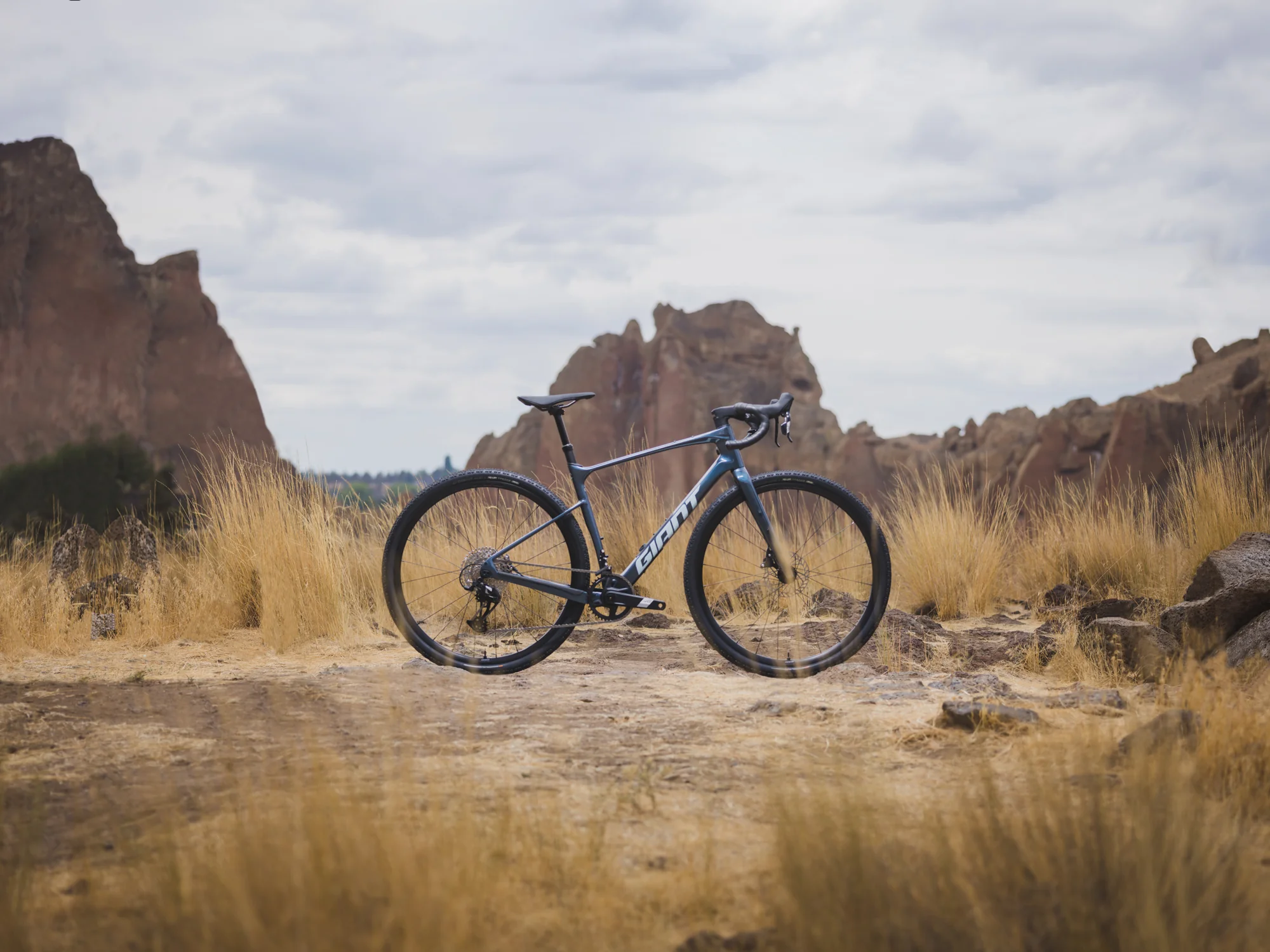 Gravel kolo Giant Revolt Advanced 1 v barvě Ocean Twilight stojí na prašné stezce mezi skalami a suchou trávou