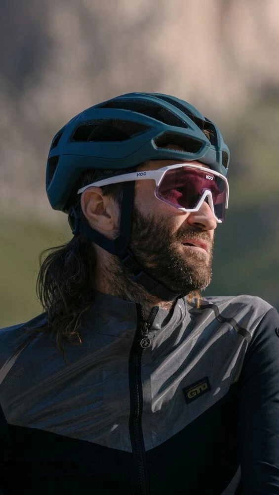 Vousatý jezdec v helmě KASK Protone Icon a sportovních brýlích, pohled z profilu před rozmazanou skálou