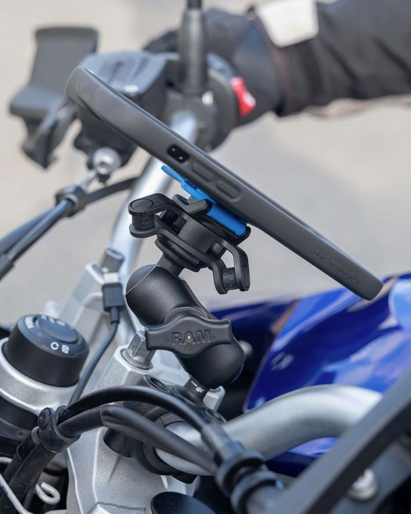 Telefon v pouzdře Quad Lock na adaptéru s tlumičem vibrací v rameni RAM u řídítek motocyklu, za ním ruka jezdce v rukavici (pouzdro, tlumič vibrací a rameno se prodávají zvlášť)