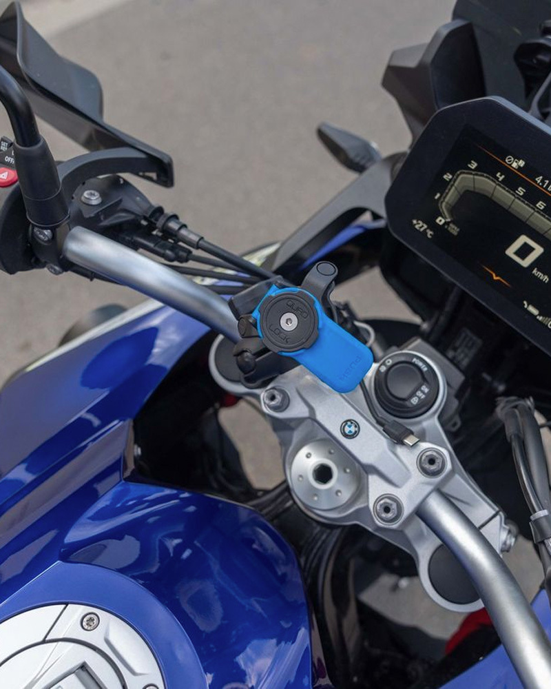 Kulový adaptér Quad Lock s modrou páčkou u řídítek modrého motocyklu, uchycený v rameni (prodává se zvlášť)