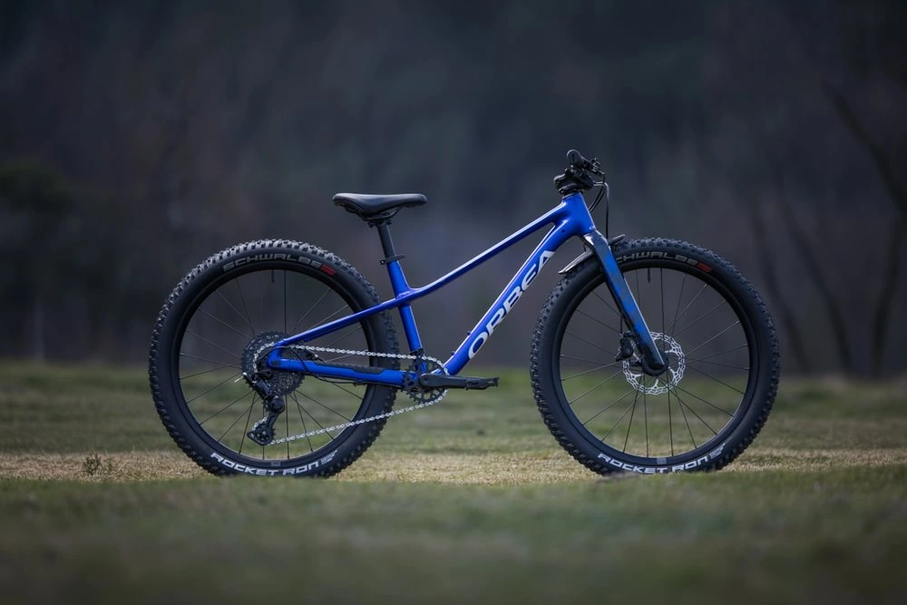 Modré dětské horské kolo Orbea Kimu 24 H10 z boku na trávě, plášť Schwalbe Rocket Ron