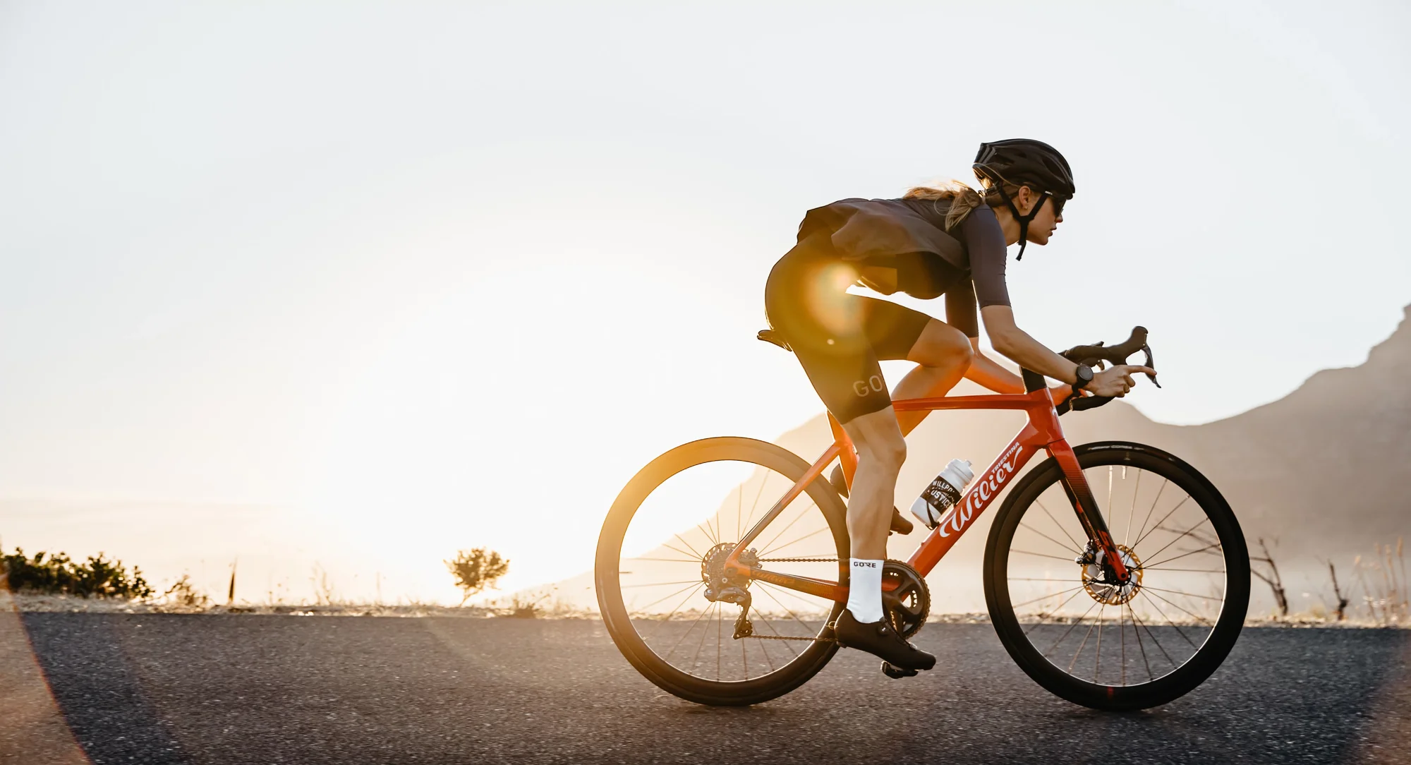 Cyklistka na silničním kole Wilier Cento10 SL při západu slunce na silnici pod horami