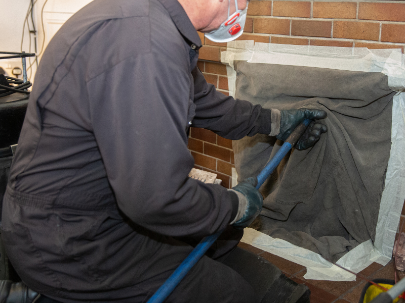 Muž čistí kouřovod krbu a výměník krbových kamen pomocí kartáče a ochranných rukavic.