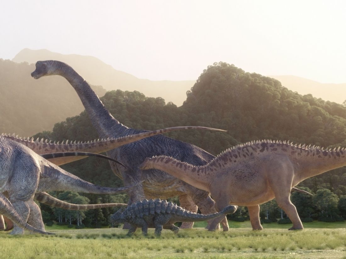 Skupina dinosaurů kráčí pravěkou krajinou s vegetací a horami v pozadí.