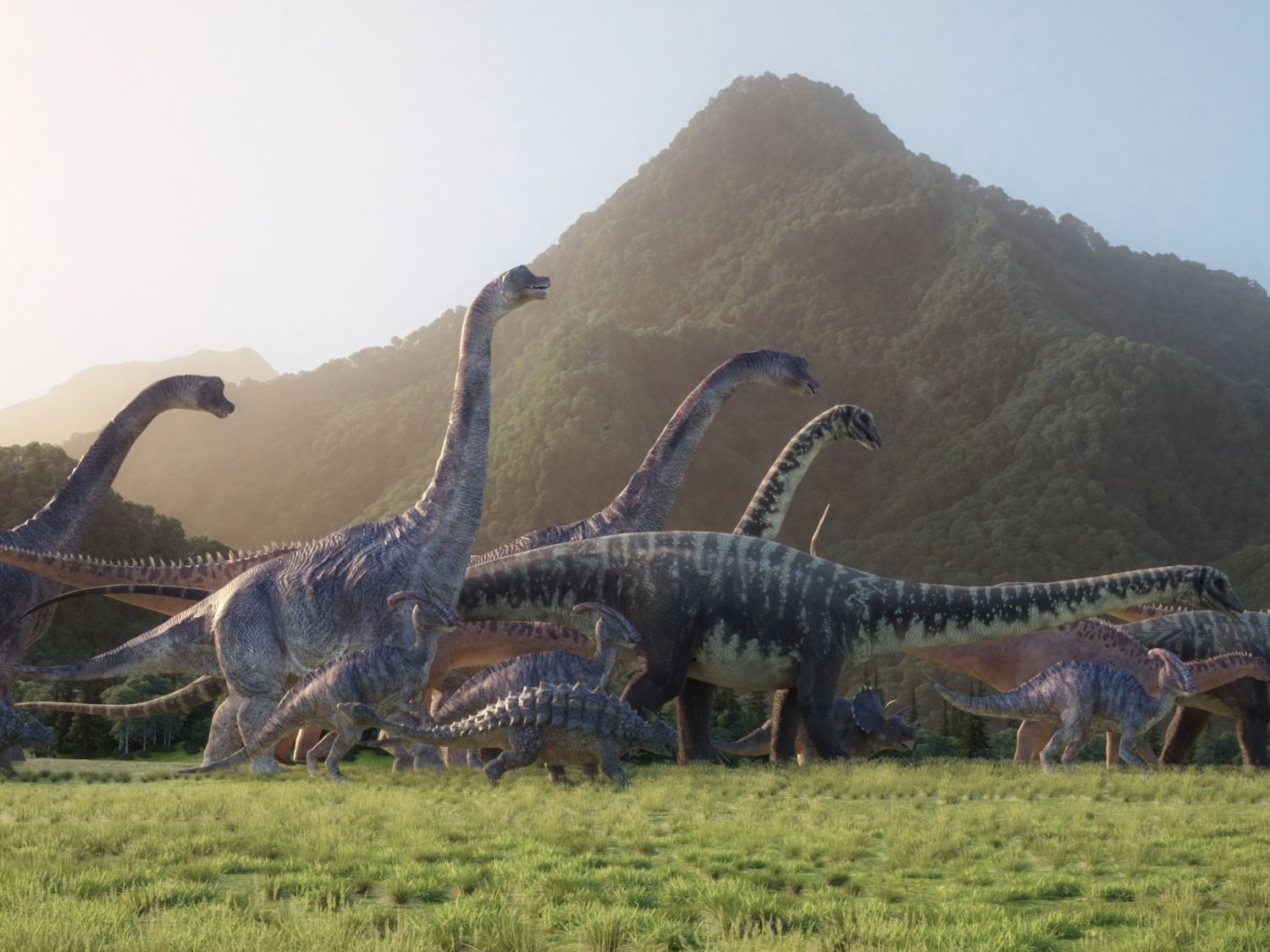 Různí dinosauři v pohybu procházejí zelenou pravěkou krajinou s horami v pozadí.