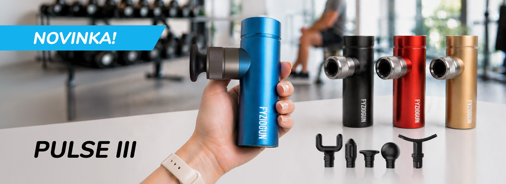 Promo banner e-shopu na novú mini masážnu pištoľ FyzioGun PULSE III. Ruka v popredí drží modrú verziu pištole s nasadeným plochým nadstavcom. Na bielej ploche vedľa sú zobrazené ďalšie tri farebné varianty (čierna, červená, zlatá) a sada piatich čiernych