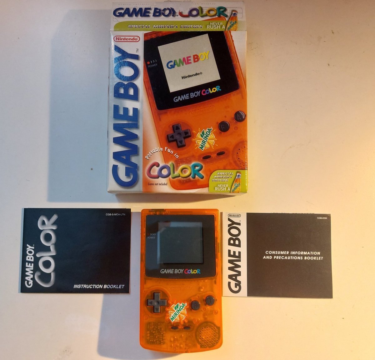Oranžový průhledný Game Boy Color s logem Mirinda mezi návodem a informační brožurou Nintendo, nad ním krabice Special Mirinda Edition