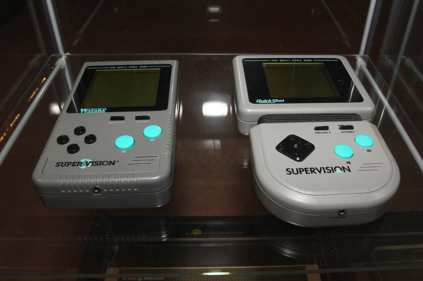 Dvě konzole Supervision ve vitríně helsinského muzea vedle sebe — vlevo plochá Watara SUPER VISION se čtyřmi kulatými tlačítky, vpravo sklopná QuickShot SUPERVISION s D-padem