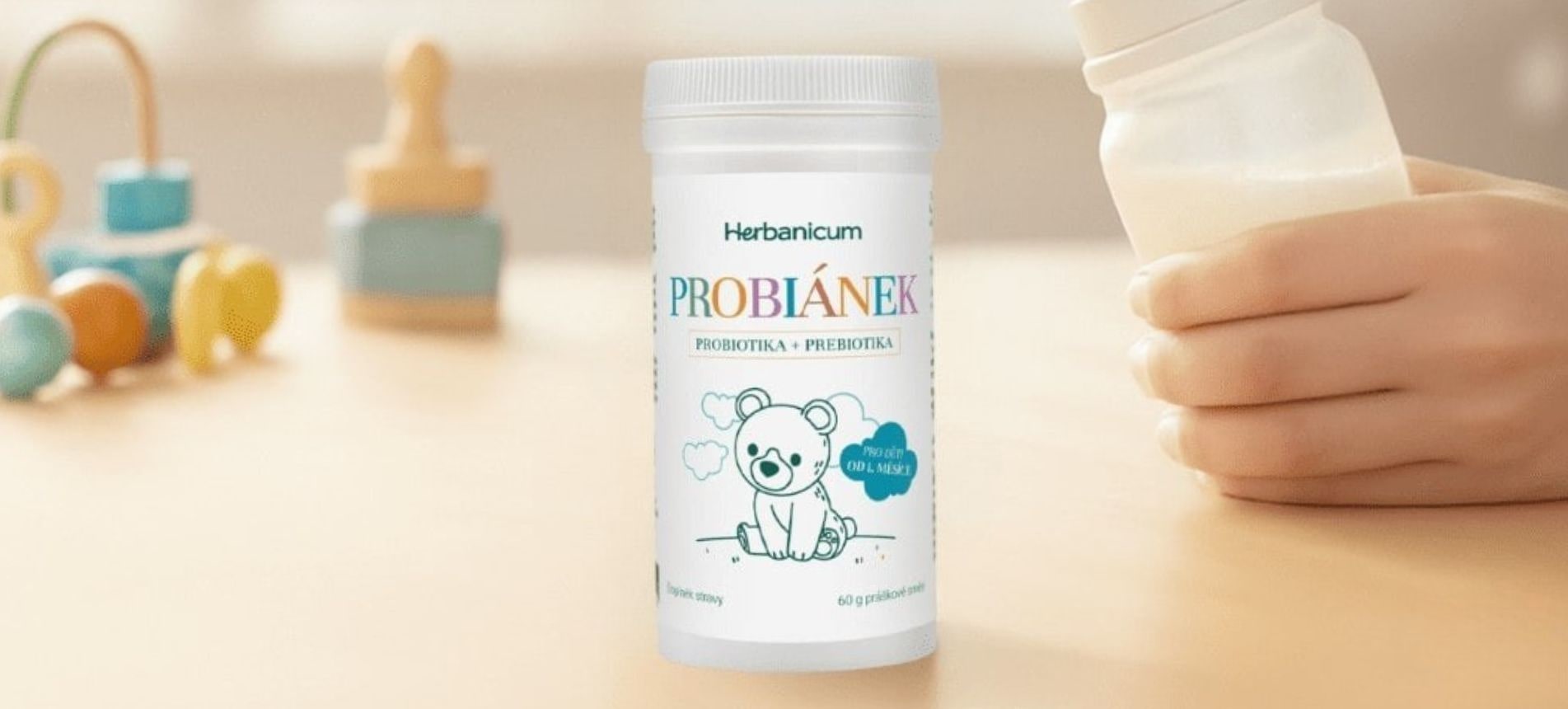 Ruka držící kojeneckou láhev s mlékem. Na stole je viditelné balení Probiánku od Herbanicum. Vizuál zdůrazňuje snadnou aplikaci pro děti 0–3 roky.