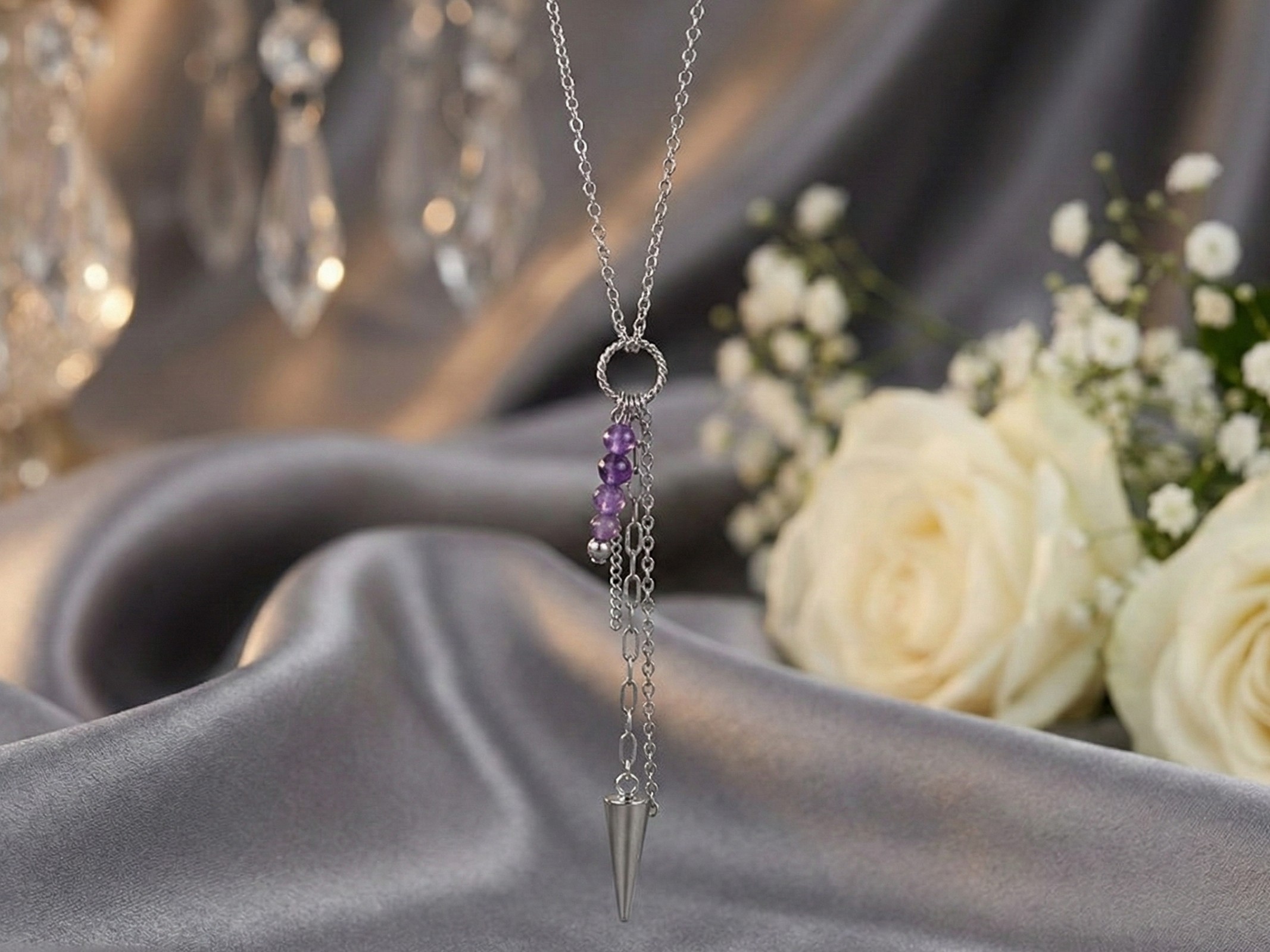Detailný pohľad na náhrdelník z chirurgickej ocele s ametystom a oceľovým hrotom, položený na saténe s kvetinovým pozadím. Elegantný a harmonický šperk.